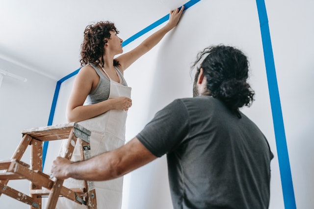 A person standing on a ladder painting a wall while another person holds it