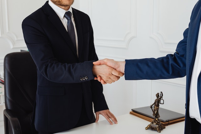 two people in suits shaking hands across a table with a status of lady justice in the background